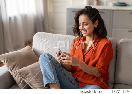 Smiling young woman browsing cellphone on sofa Smiling young woman browsing cellphone on sofa 112721119