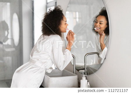 Young black lady brushes teeth in front of bathroom mirror 112721137