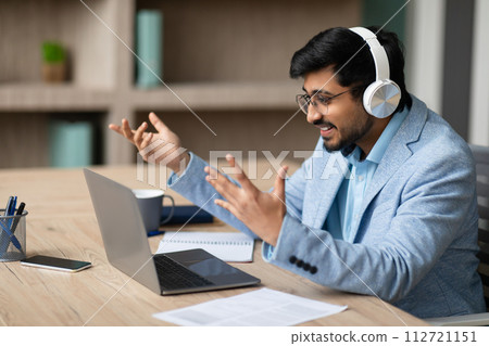 Hindu businessman wearing headphones communicating through video call in office Hindu businessman wearing headphones communicating through video call in office 112721151
