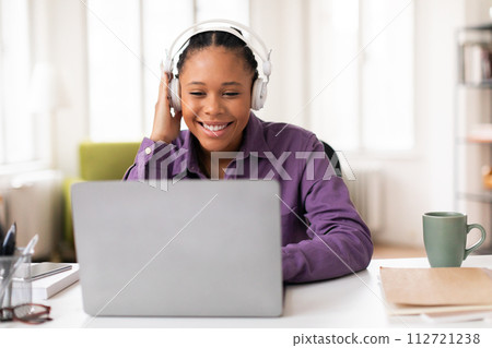 Happy black lady student looking at laptop and touching headphones 112721238
