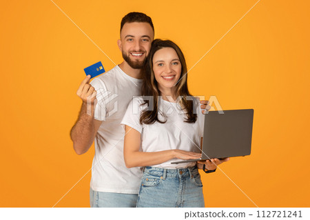 Happy couple engaged in online shopping, with the man holding a credit card and the woman holding a laptop 112721241