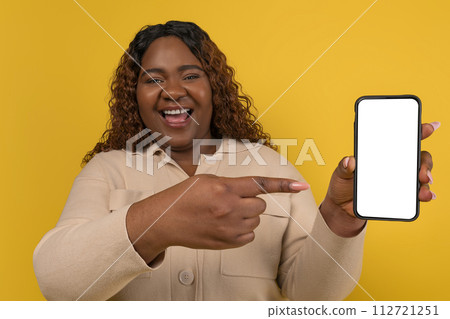 Positive chubby african american lady pointing at phone in hand 112721251