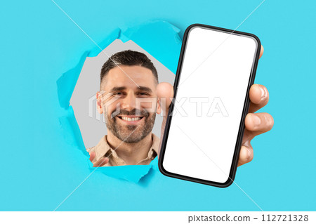 Smiling bearded man looking through a ripped cyan paper, extending a hand to present a smartphone 112721328