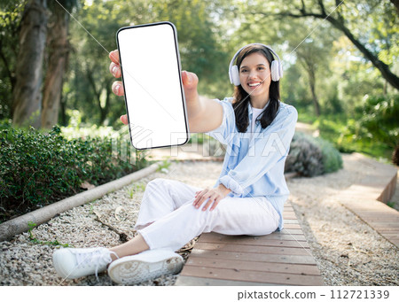 A young woman with headphones smiles brightly, showcasing her smartphone on a sunny park trail 112721339