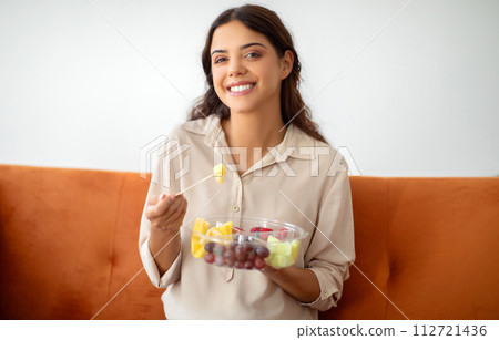 Young beautiful woman eating fruit salad while sitting on couch at home 112721436