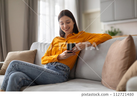 Smiling young woman browsing phone, relaxed on sofa 112721480