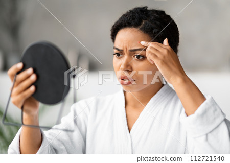 Concerned black woman looking in handheld mirror at pimple on her forehead 112721540