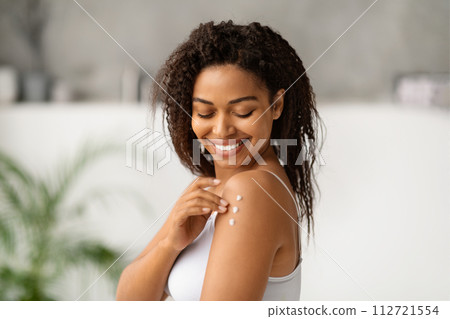 Joyful black woman smiling while applying body lotion to her shoulder Joyful black woman smiling while applying body lotion to her shoulder 112721554