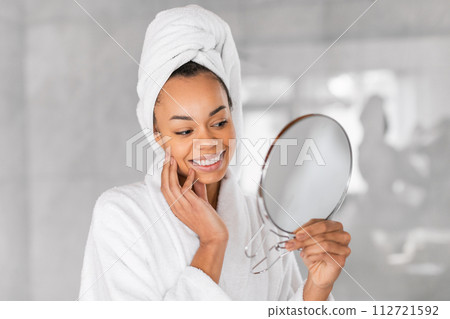 Black millennial woman in bathrobe holding a round mirror indoor Black millennial woman in bathrobe holding a round mirror indoor 112721592