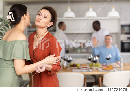 Women greet each other with kiss on cheek before festive dinner. Men shaking hands in background Women greet each other with kiss on cheek before festive dinner. Men shaking hands in background 112721676