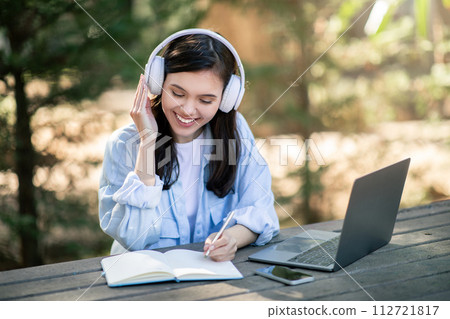 A joyful young woman enjoys her music with headphones, writing in a notebook beside her laptop 112721817
