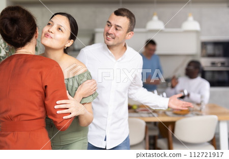 Women greet each other with kiss on cheek before festive dinner. Men shaking hands in background 112721917