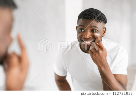 Cheerful millennial black guy applying moisturizer on face in bathroom 112721932