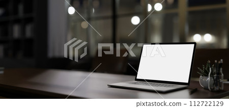 A modern dark office at night features a white-screen laptop computer mockup on a table. A modern dark office at night features a white-screen laptop computer mockup on a table. 112722129