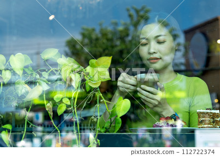 Asian woman enjoying her coffee in a cafe, viewed through a window with reflections of foliage Asian woman enjoying her coffee in a cafe, viewed through a window with reflections of foliage 112722374