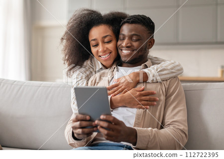 Loving millennial African American couple enjoys websurfing on tablet indoors Loving millennial African American couple enjoys websurfing on tablet indoors 112722395