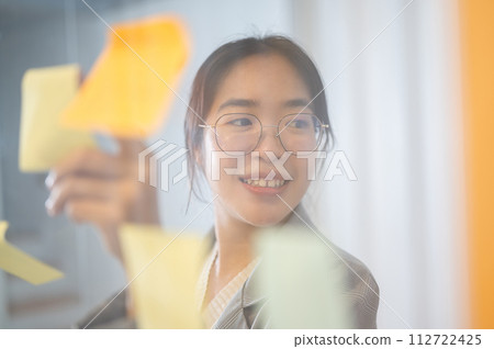A positive Asian businesswoman brainstorming with her team, reading ideas on sticky notes. 112722425