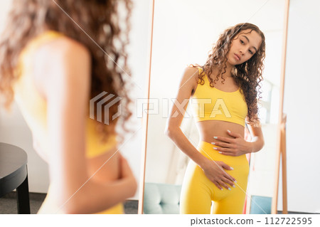 Upset teen girl in fitwear touches belly near mirror indoor Upset teen girl in fitwear touches belly near mirror indoor 112722595