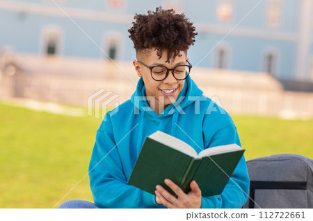 Happy african american student guy in eyeglasses reading book outside 112722661