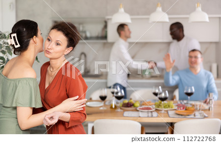 Women greet each other with kiss on cheek before festive dinner. Men shaking hands in background 112722768