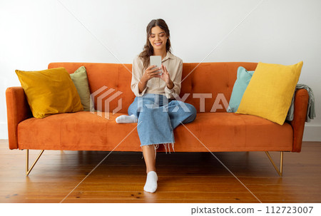 Content young woman in casual wear smiling while using smartphone at home Content young woman in casual wear smiling while using smartphone at home 112723007