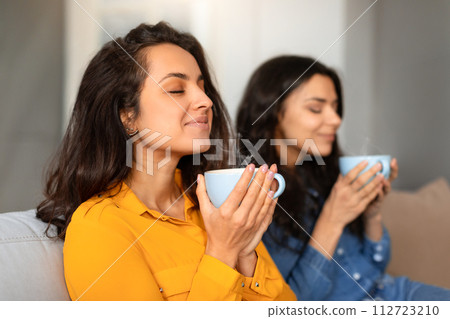 Two young women enjoying aroma of coffee holding cups indoors 112723210