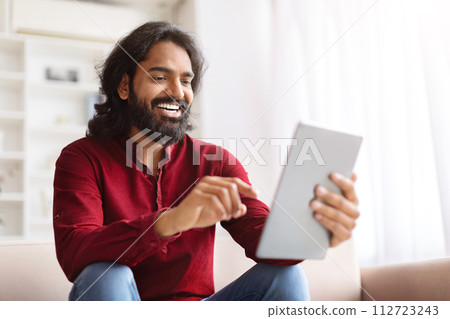 Cool millennial indian guy sitting on couch, using digital tablet Cool millennial indian guy sitting on couch, using digital tablet 112723243