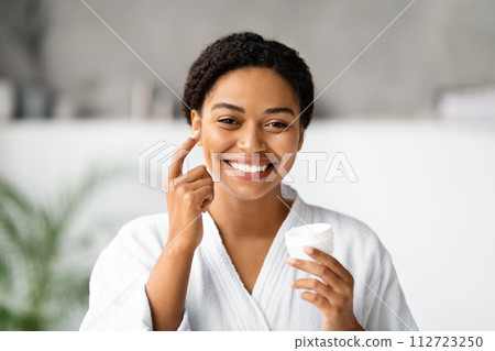 Cheerful black woman applying moisturizing cream on her cheek 112723250