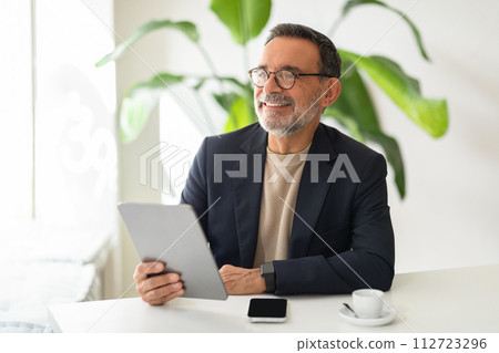 A contented senior businessman in a stylish navy blazer and beige turtleneck smiles while using a digital tablet 112723296