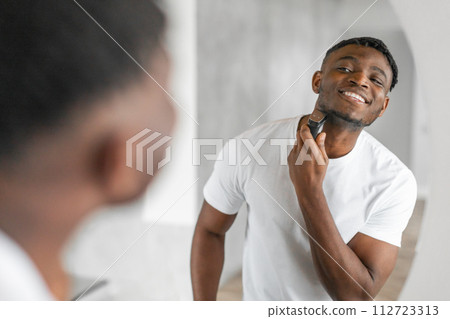 Happy African American man shaving with an electric razor indoor Happy African American man shaving with an electric razor indoor 112723313