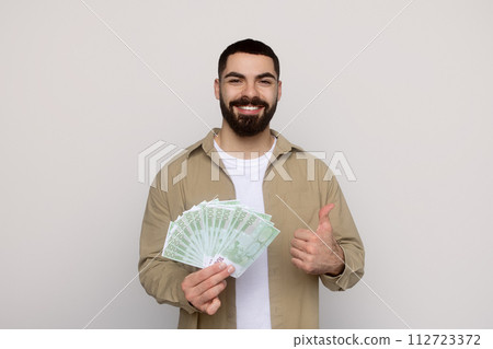 Smiling man in a beige shirt holds a fan of 100-euro bills in one hand and gives a thumbs up 112723372