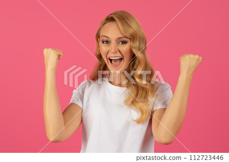 blonde woman celebrating success shaking clenched fists on pink backdrop 112723446