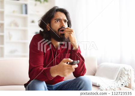 Shocked eastern guy with remote control sitting on couch 112723535