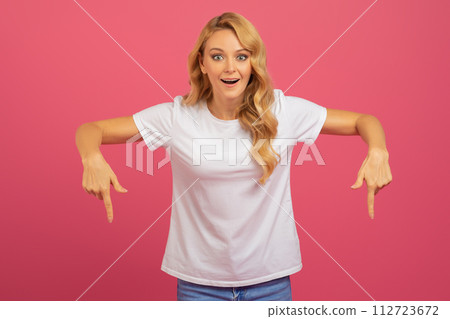 Blonde woman pointing fingers downward with excitement over pink background 112723672