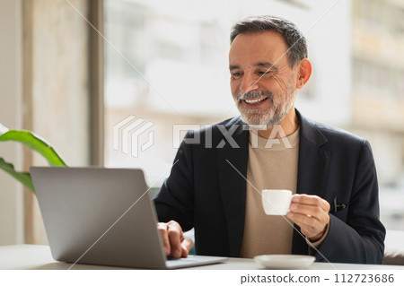 Cheerful senior man with a beard enjoying a cup of coffee while working on a laptop Cheerful senior man with a beard enjoying a cup of coffee while working on a laptop 112723686