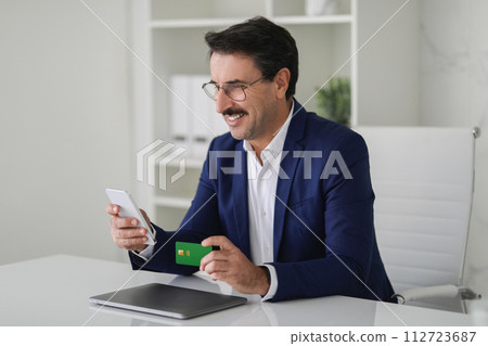 Joyous businessman in a blue suit engaging with his mobile phone while holding a debit card Joyous businessman in a blue suit engaging with his mobile phone while holding a debit card 112723687