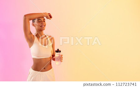 Relaxed fit woman enjoying a break during her workout, holding a water bottle with eyes closed 112723741