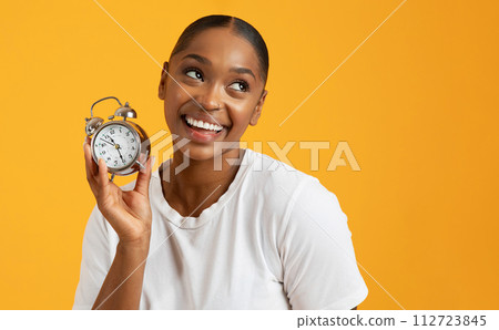 Cheerful black millennial woman holding alarm clock on yellow background Cheerful black millennial woman holding alarm clock on yellow background 112723845