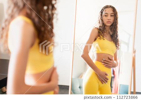 Troubled lady in workout attire contemplates reflection touching belly indoor 112723866