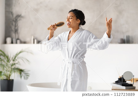 Happy young black woman holding hairbrush like microphone while singing in bathroom Happy young black woman holding hairbrush like microphone while singing in bathroom 112723889
