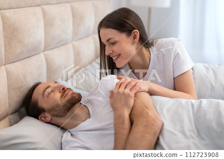 Affectionate woman smiling at her resting partner in bed Affectionate woman smiling at her resting partner in bed 112723908