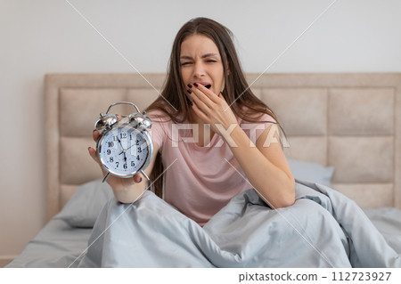 Sleepy woman in bed holding an alarm clock, yawning with tiredness Sleepy woman in bed holding an alarm clock, yawning with tiredness 112723927