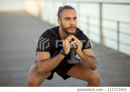 Athlete man performing squats holding dumbbell weights working out outside 112723942