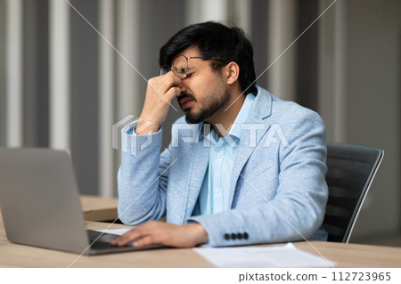 Indian businessman experiencing eye strain sitting tired in office Indian businessman experiencing eye strain sitting tired in office 112723965