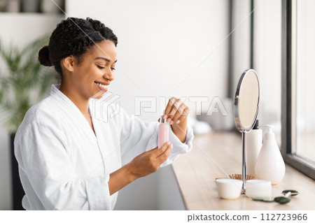 Smiling black woman holding bottle of facial serum, ready to apply skincare 112723966