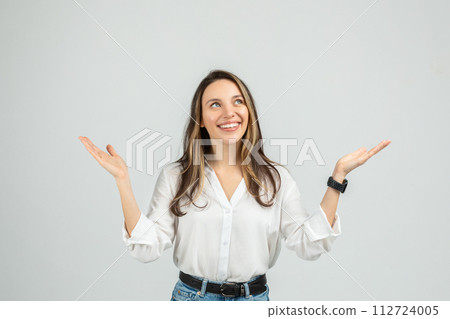 An upbeat young woman in a white blouse and blue jeans is raising her hands, palms up 112724005