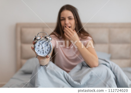 Tired woman in bed with alarm clock, yawning early in the morning 112724020
