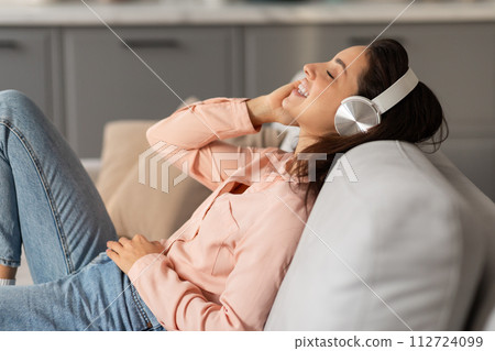 Joyful woman lying on couch with headphones, feeling relaxed 112724099