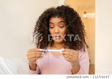 Shocked black woman checking pregnancy test at home, feeling surprised with result 112724247