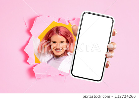 Smiling excited young caucasian lady showing mobile smartphone with empty screen through hole in paper 112724266